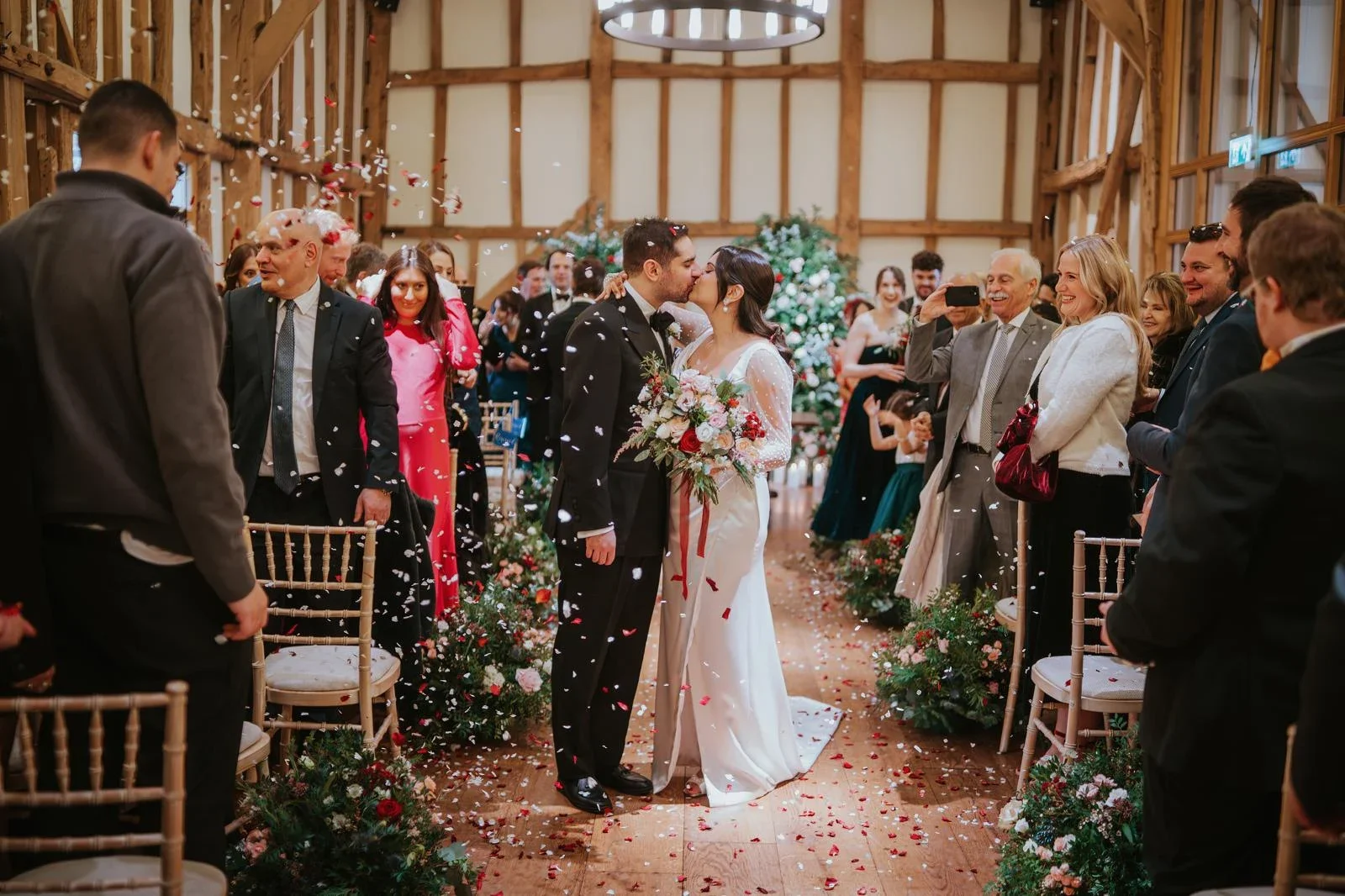 Bride and groom kissing under a shower of confetti as they walk back down the aisle of a timber-framed barn