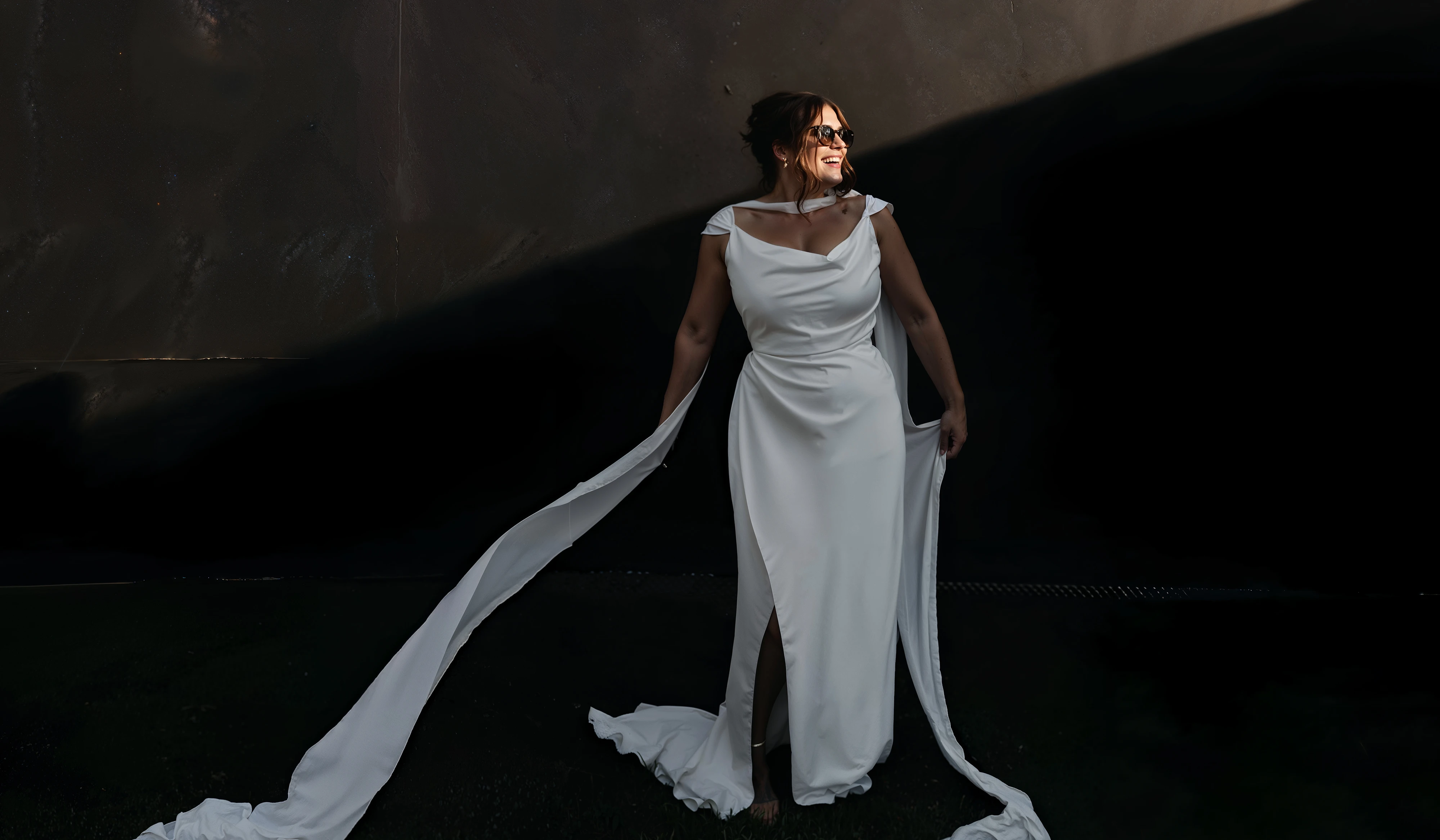 Bride in a draped satin cowl-neck wedding dress with long flowing cape sleeves, lit against a dark wall