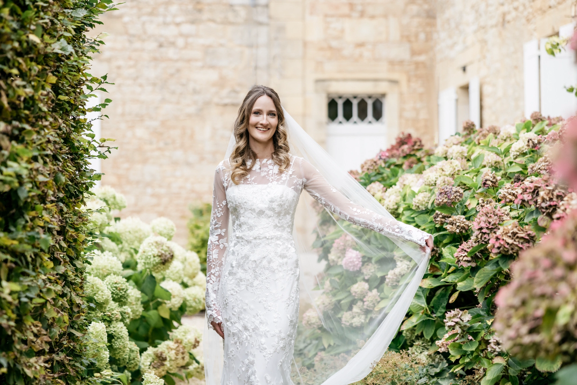 Custom Italian silk wedding gown with lace sleeves, bride holding her veil among the hydrangeas