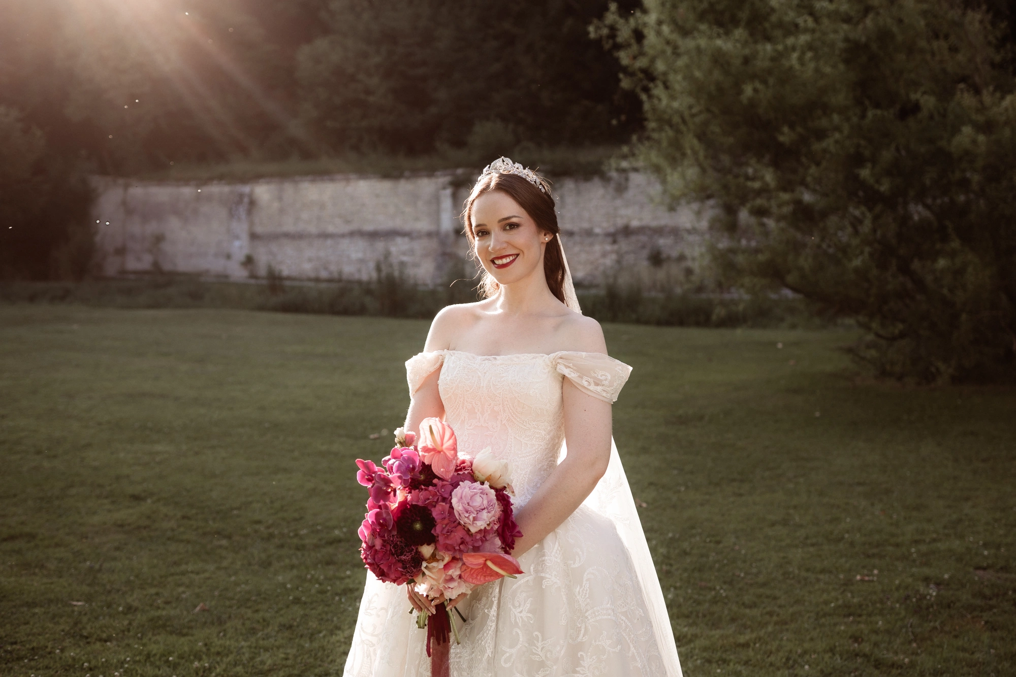 Custom ivory lace ballgown wedding dress, front view with her bouquet in the walled garden at sunset