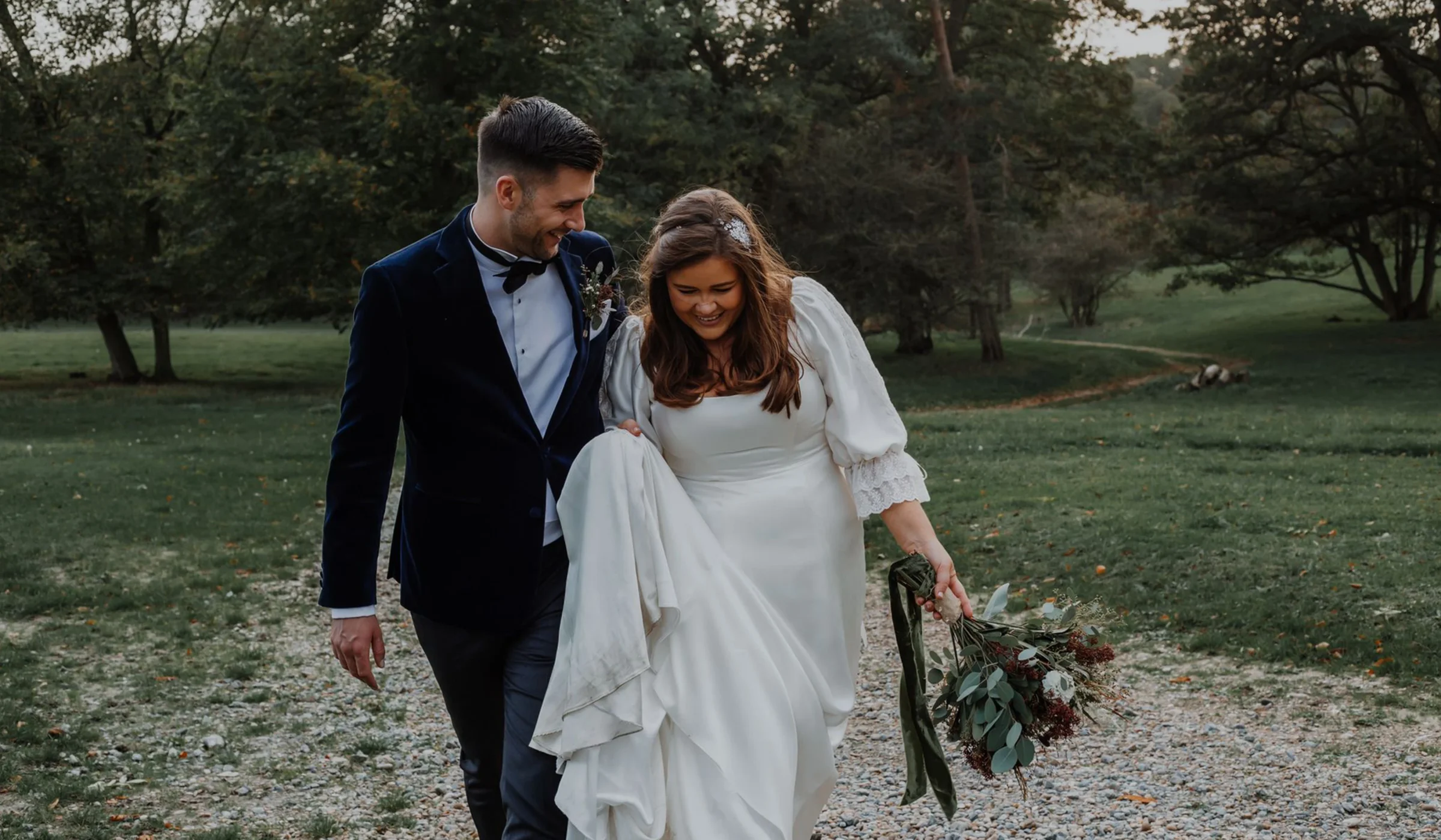 Bride in a satin wedding dress with lace-trimmed sleeves walking a woodland path with her groom in a navy velvet jacket